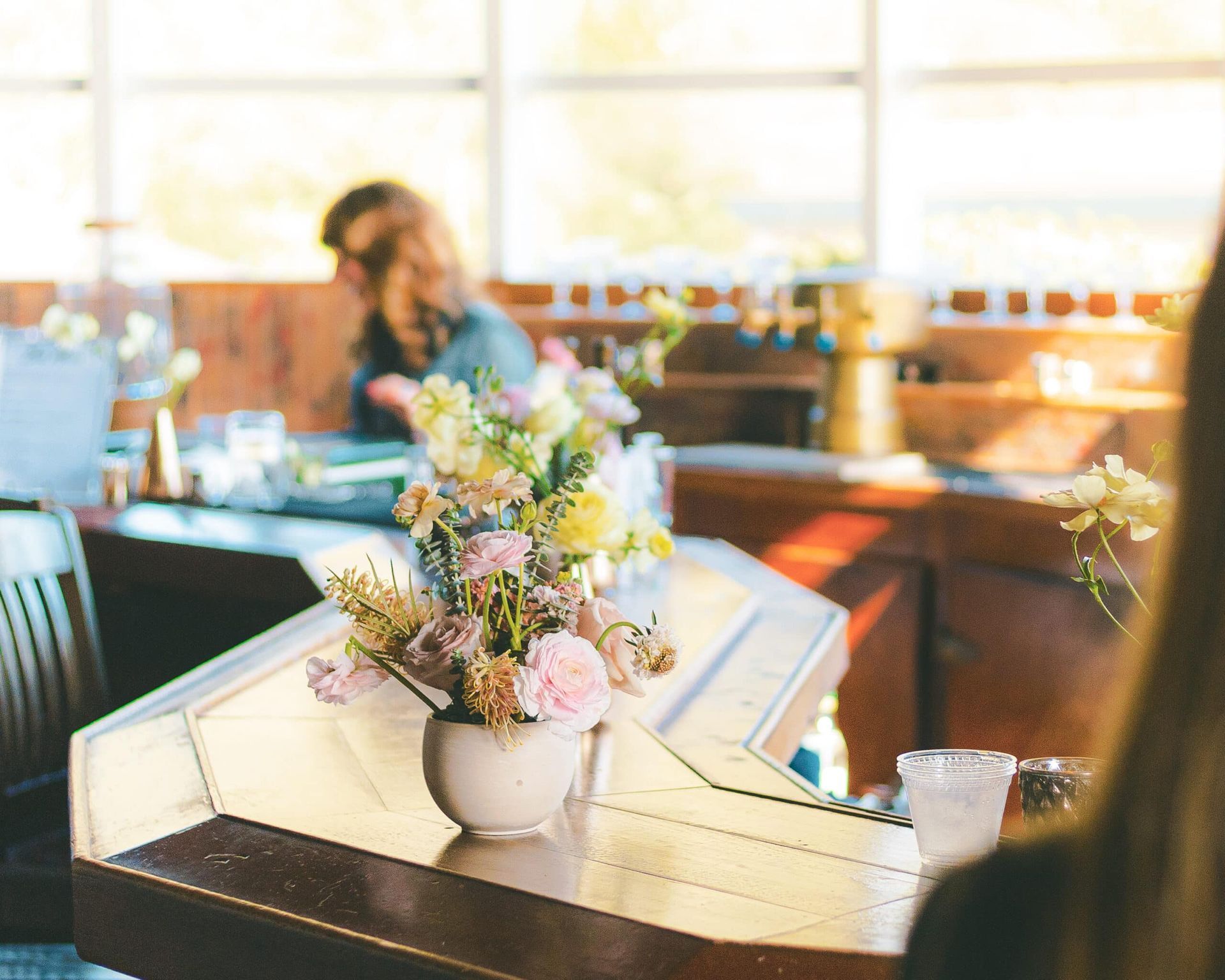 Flower arrangement on a wood table in a cafe, person blurred in background, natural light.