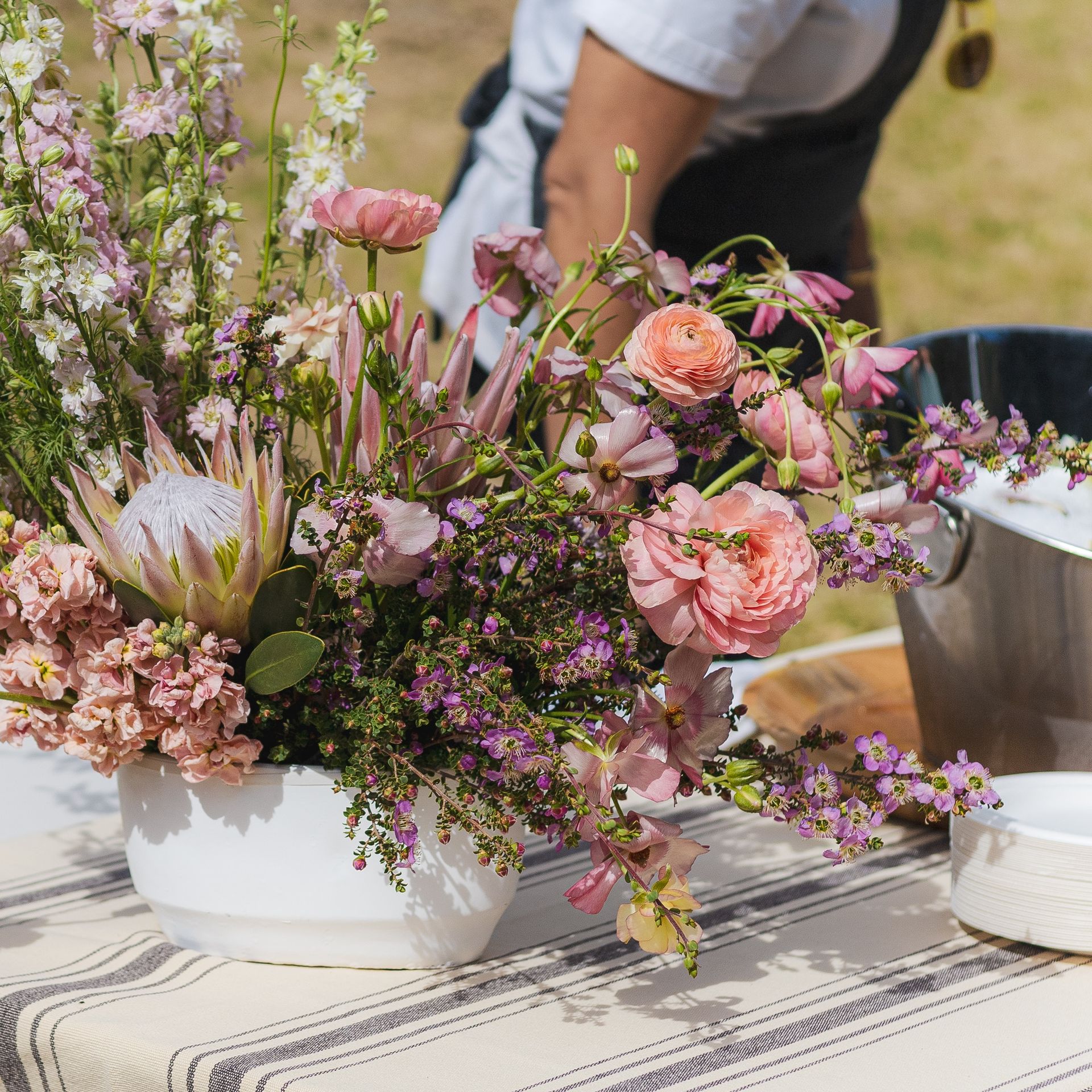 White ceramic bowl overflowing with pink and purple flowers on a striped tablecloth, with person in background.