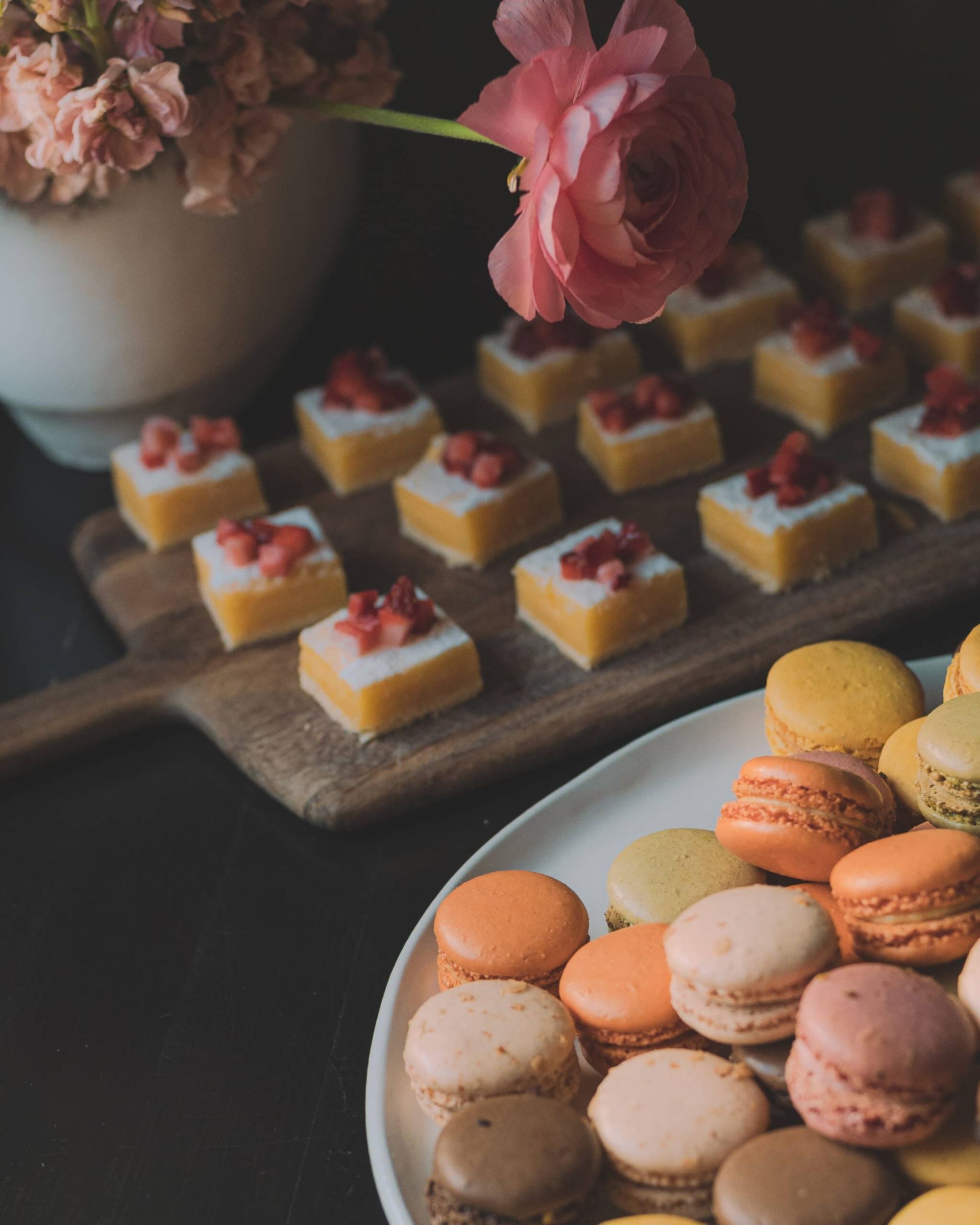 Pastries on a wooden board and a plate: small squares topped with berries and colorful macarons.
