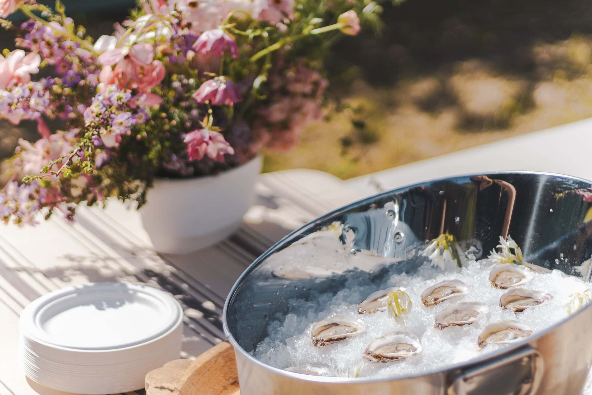 Silver bucket with oysters on ice, flowers, and white plates on a table.