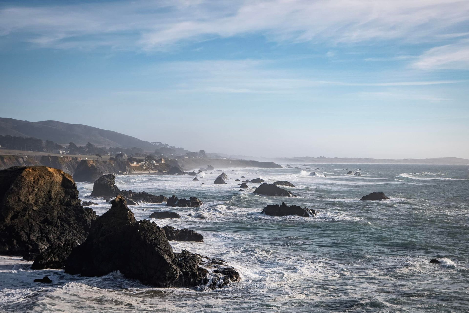 Rocky coastline under a blue sky, waves crashing against dark rocks.