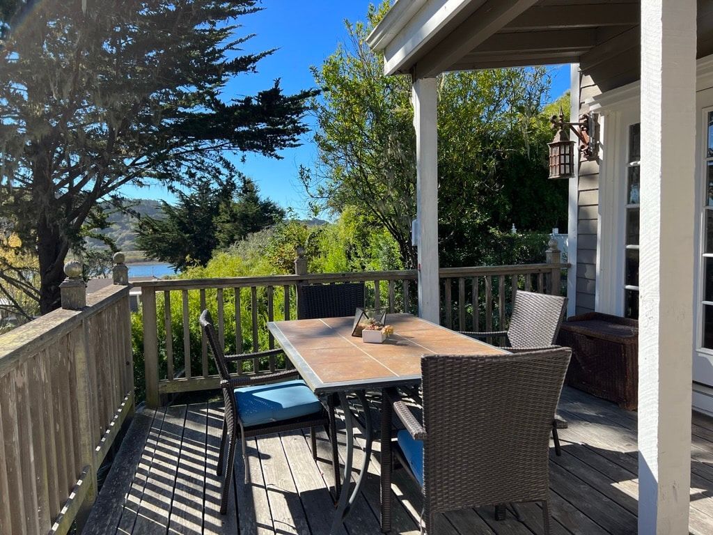 Wooden deck with table and chairs under a covered porch overlooking trees and water. Sunny day.