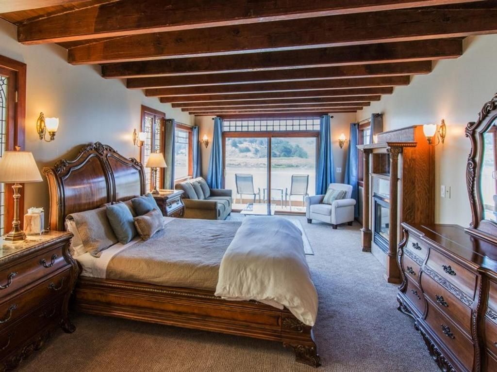 Bedroom with wooden ceiling beams, large bed, seating area, and sliding doors to a patio overlooking a lake.