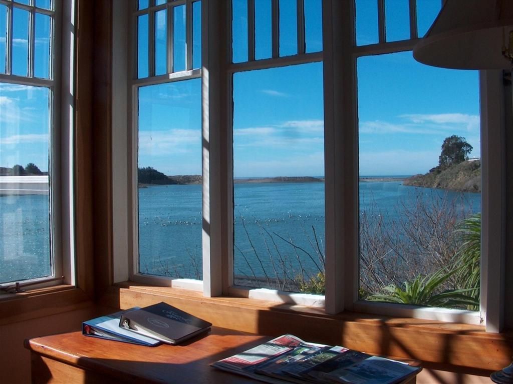 Wooden-framed windows overlooking a blue ocean view. A desk with books sits in front of the windows.