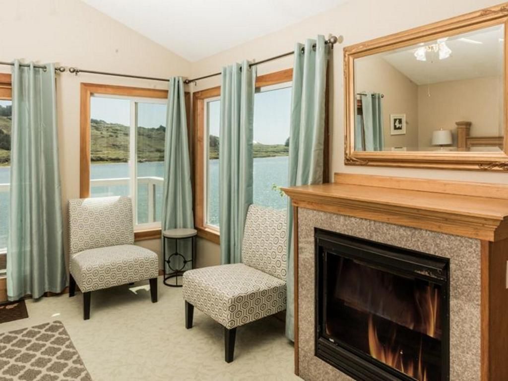 Cozy room with fireplace, chairs, and window overlooking water, light blue curtains and a large mirror.