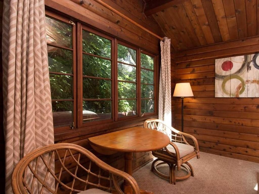 Cozy wooden room with window overlooking trees, table, two wicker chairs, lamp, and abstract art.