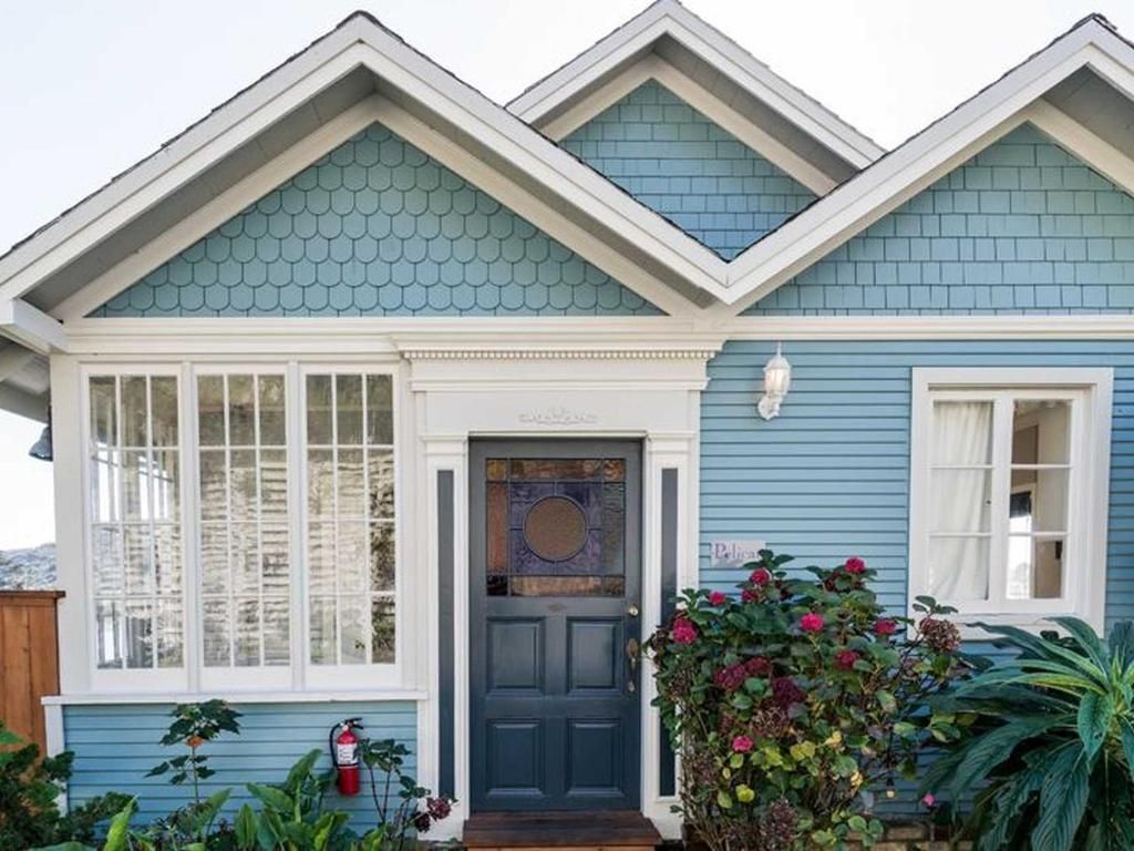 Blue cottage with white trim, teal shingles, and a dark blue door surrounded by lush greenery.
