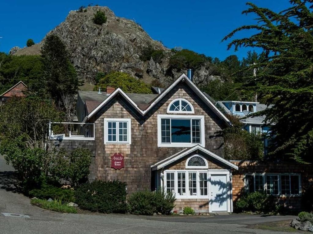 Brown shingled house with white trim. Rocky mountain in background. Blue sky, green foliage.
