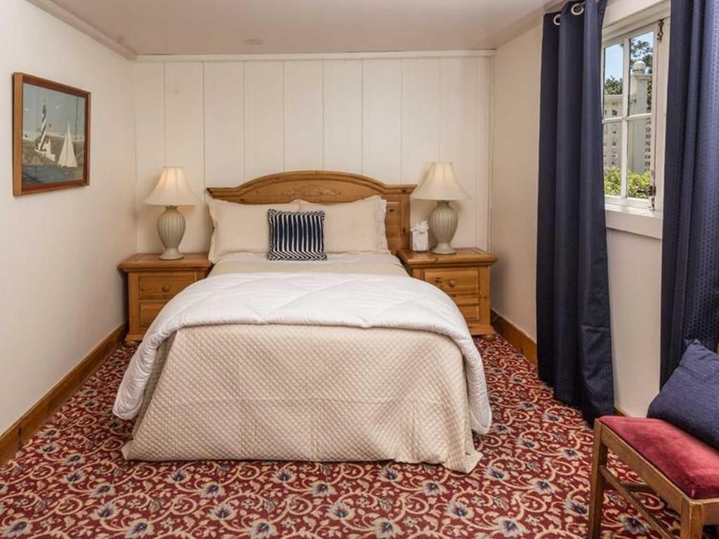 Bedroom with a bed, nightstands, lamps, a painting, patterned carpet, and a window with curtains.