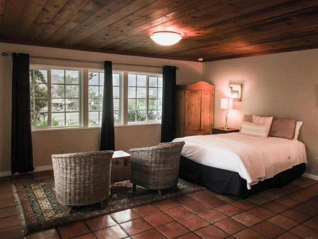 Bedroom with wooden ceiling, tiled floor, bed, wicker chairs, and windows overlooking a landscape.