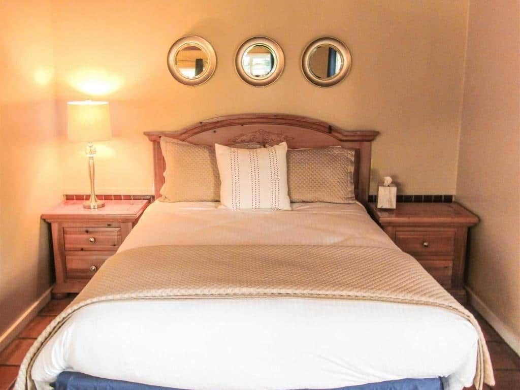 Bedroom with queen-sized bed, bedside tables, lamps, three circular mirrors above headboard, tan and cream tones.