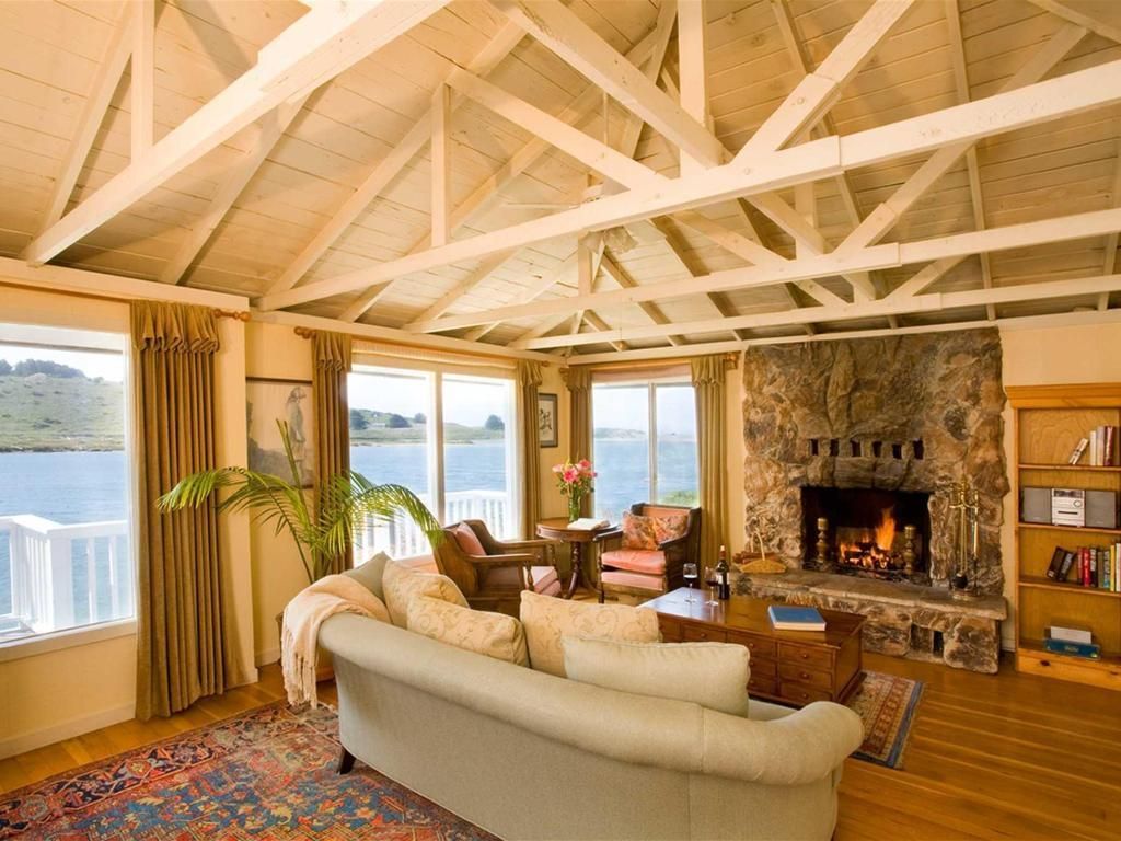 Living room with stone fireplace, wood beams, and large windows overlooking water.