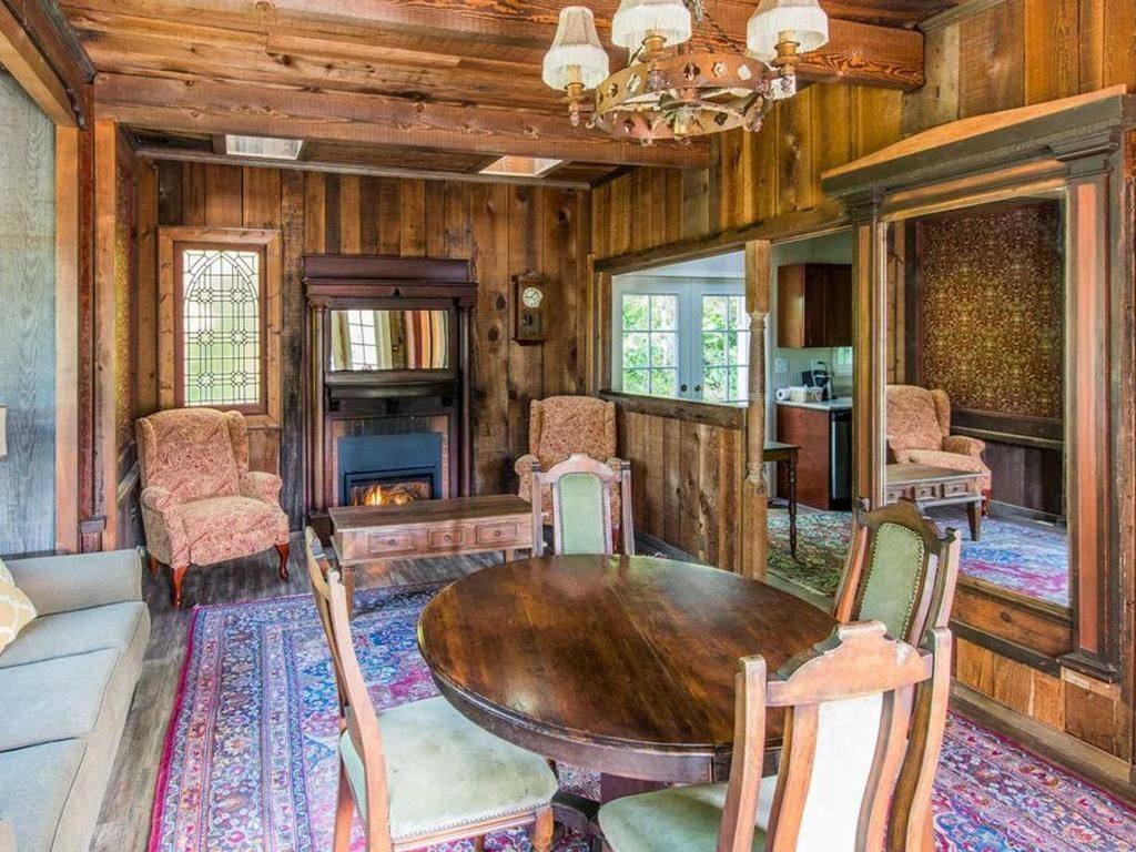 Rustic wood-paneled room with fireplace, round dining table, antique chairs, and a chandelier.