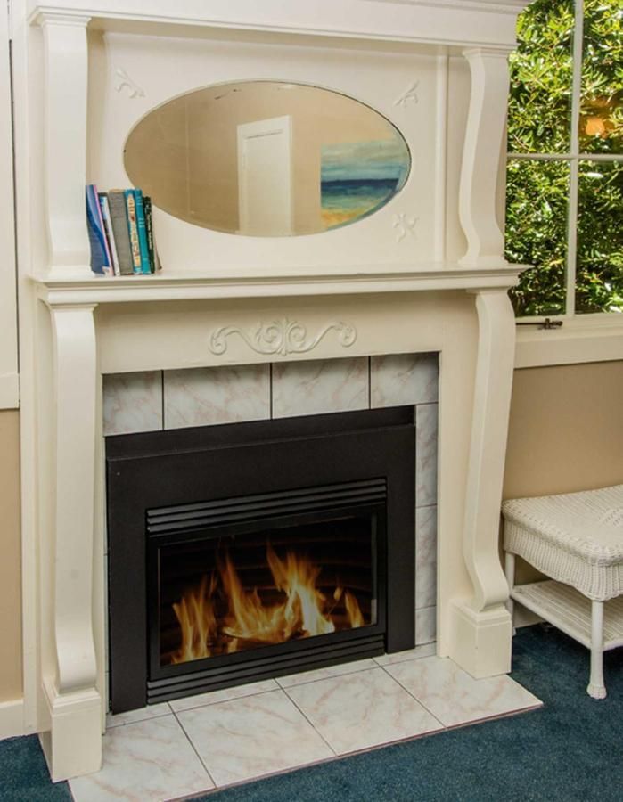 White fireplace with a lit fire, an oval mirror, and books on the mantle. A window and chair are beside it.