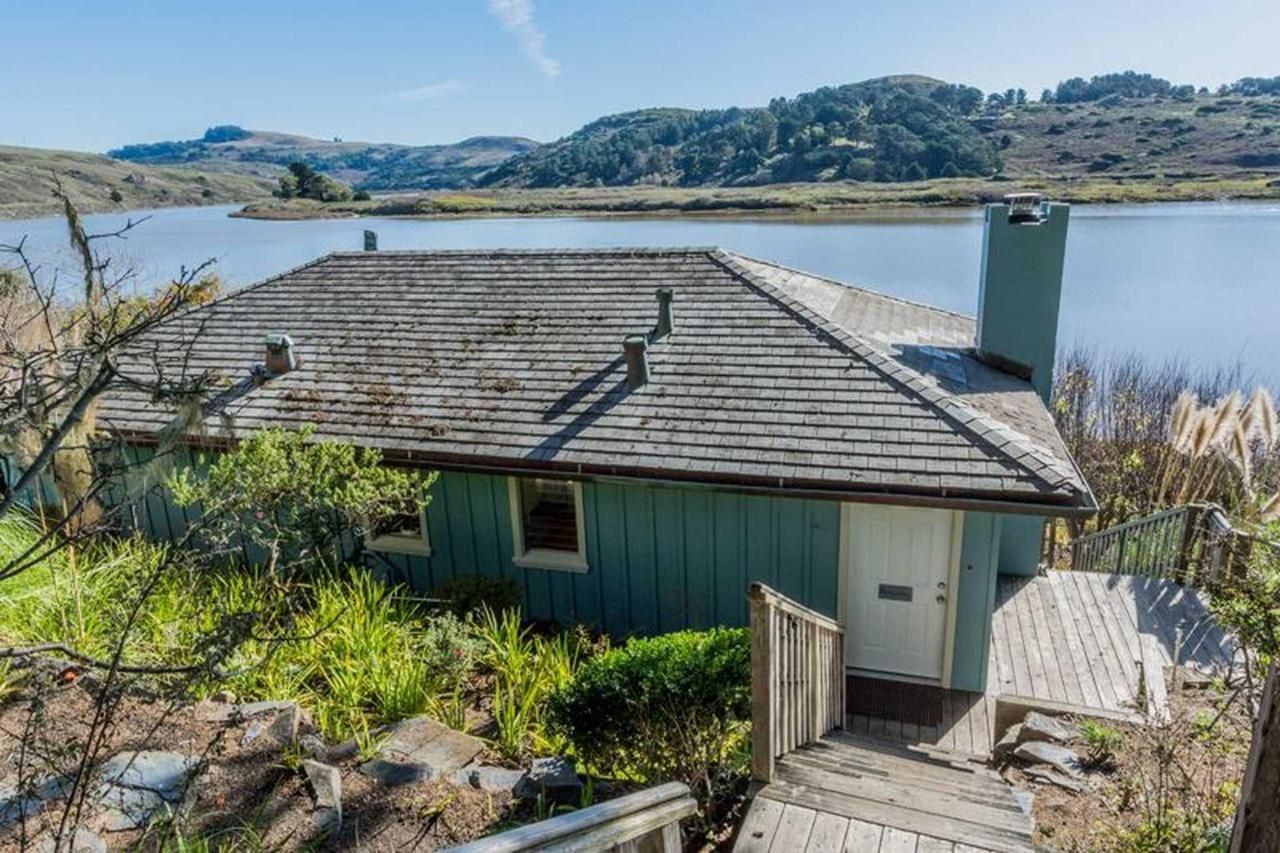Blue cabin with wooden steps, overlooking a lake and hills.