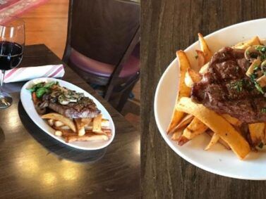 Two plates of steak frites with red wine on a wooden table.