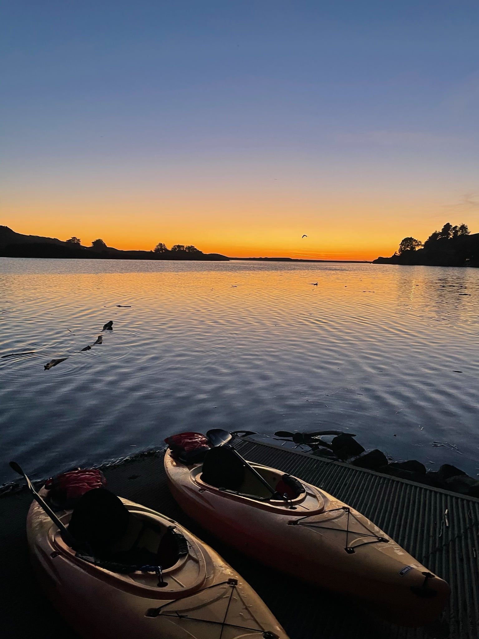 Two kayaks on a dock at sunset with orange and blue sky reflected in calm water.