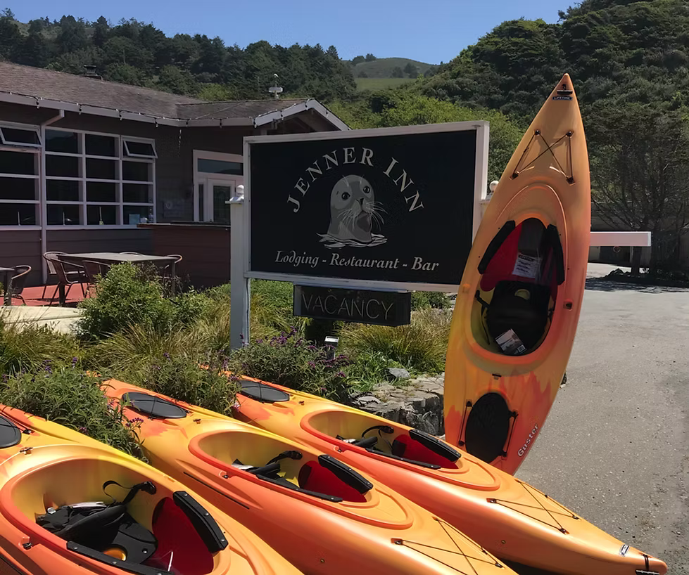 Row of orange kayaks in front of Jenner Inn sign with lodging, restaurant, bar, and vacancy details.