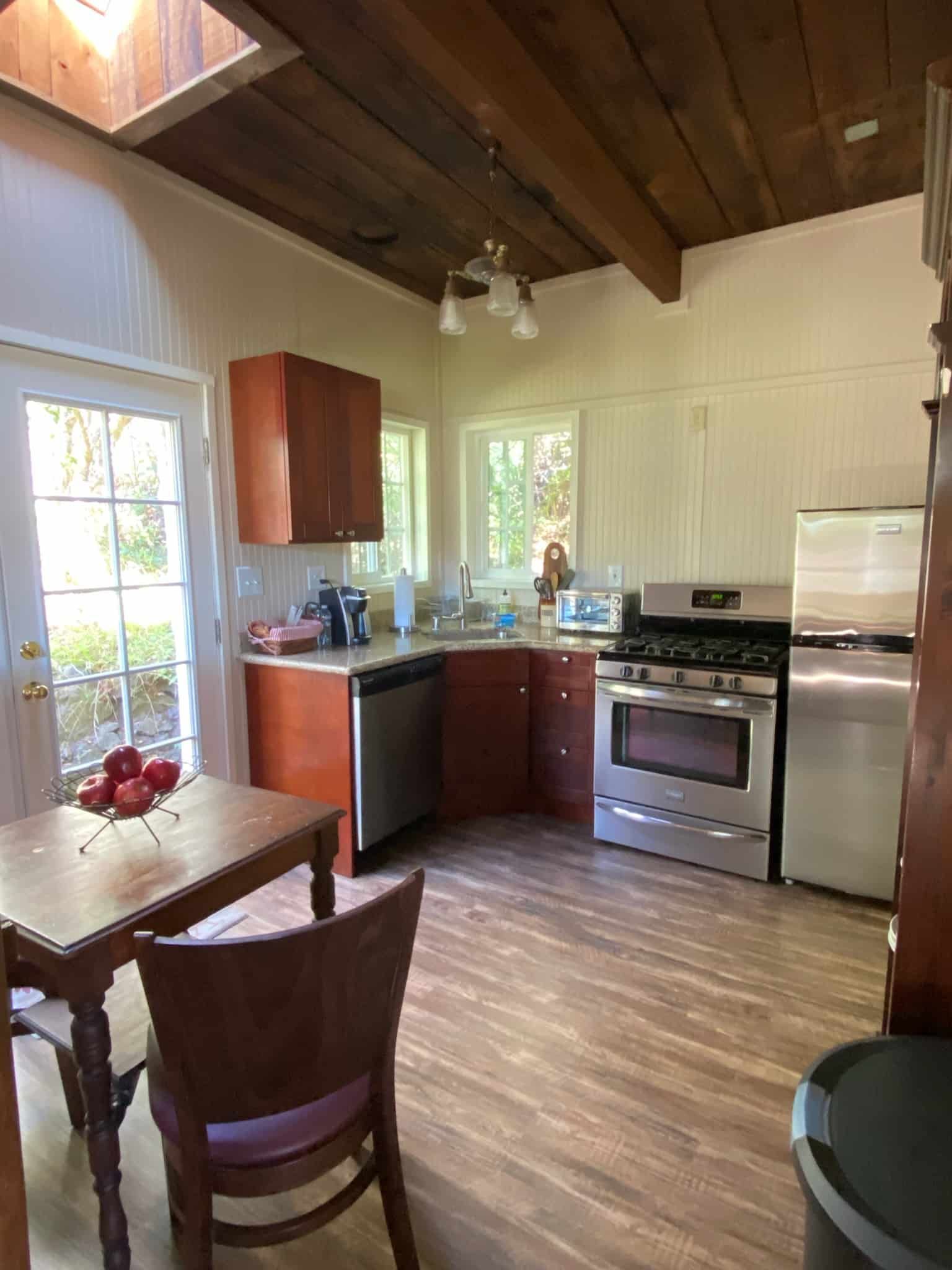 Kitchen with wood ceiling, cabinets, and appliances. Table and chair in the foreground. Door to the left.