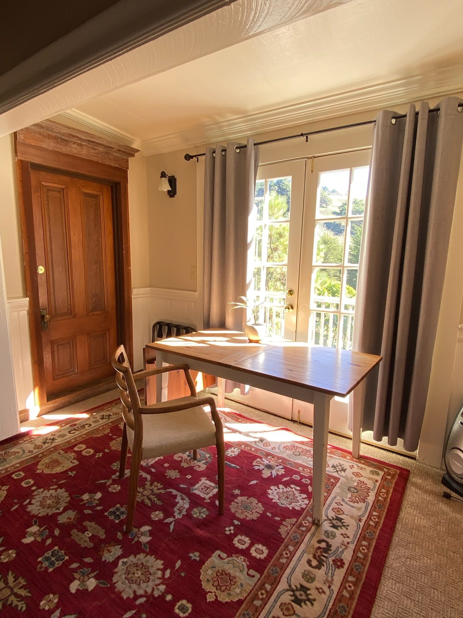 A room with a wooden door, desk and chair on a red patterned rug, next to an open door with curtains.
