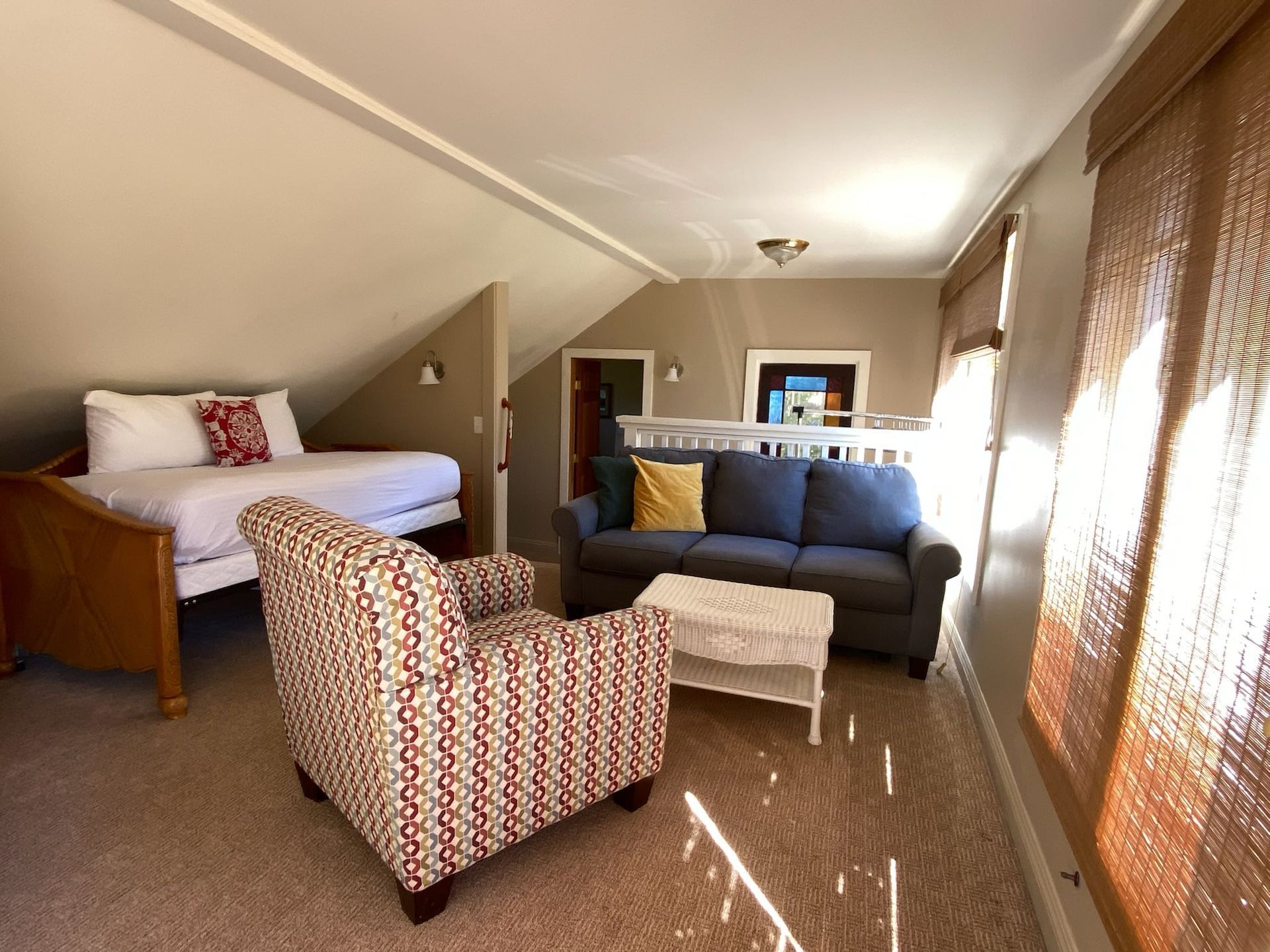 Loft room with bed, sofa, patterned armchair, and woven window shades.