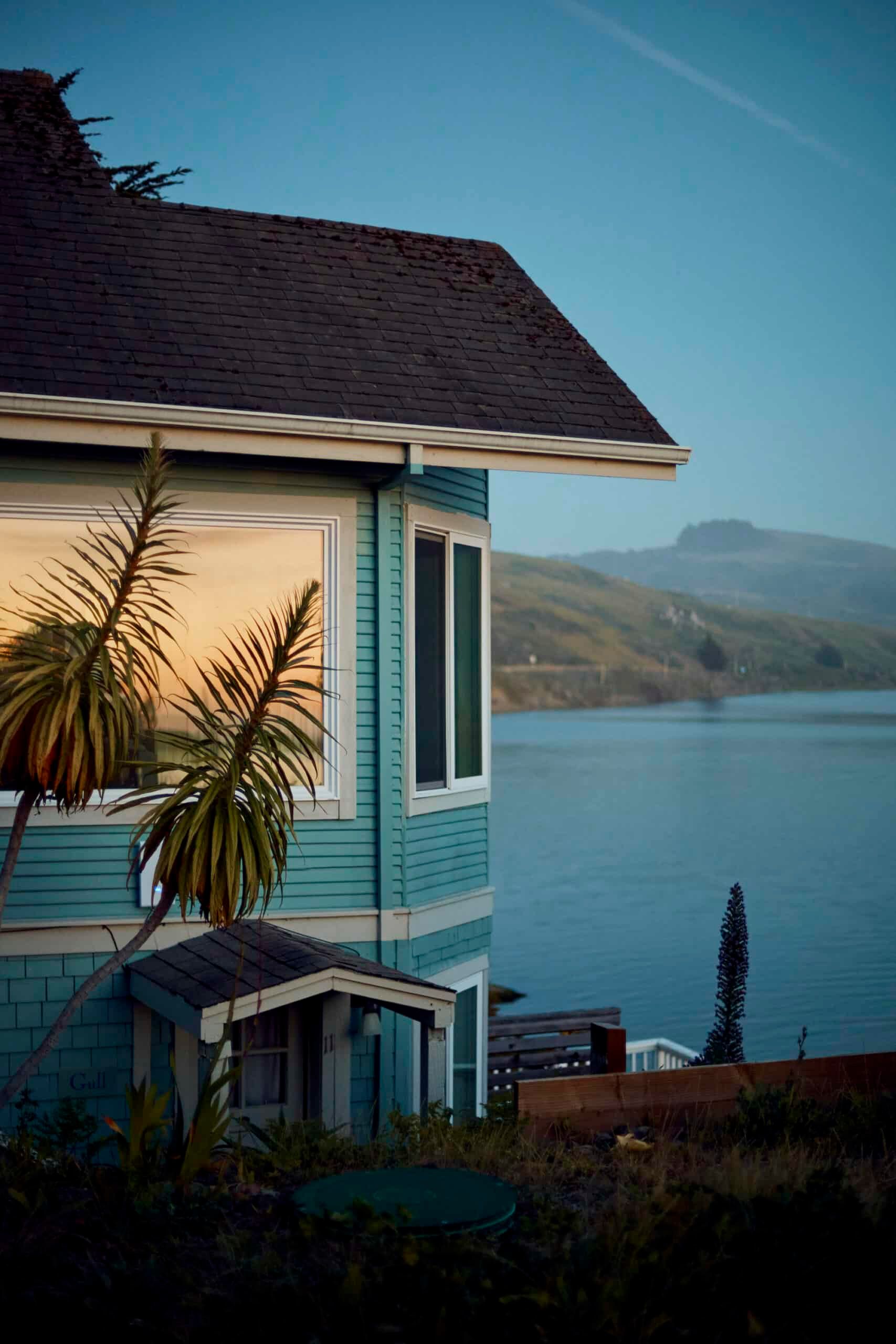 Blue house overlooking a calm body of water, with hills in the background.