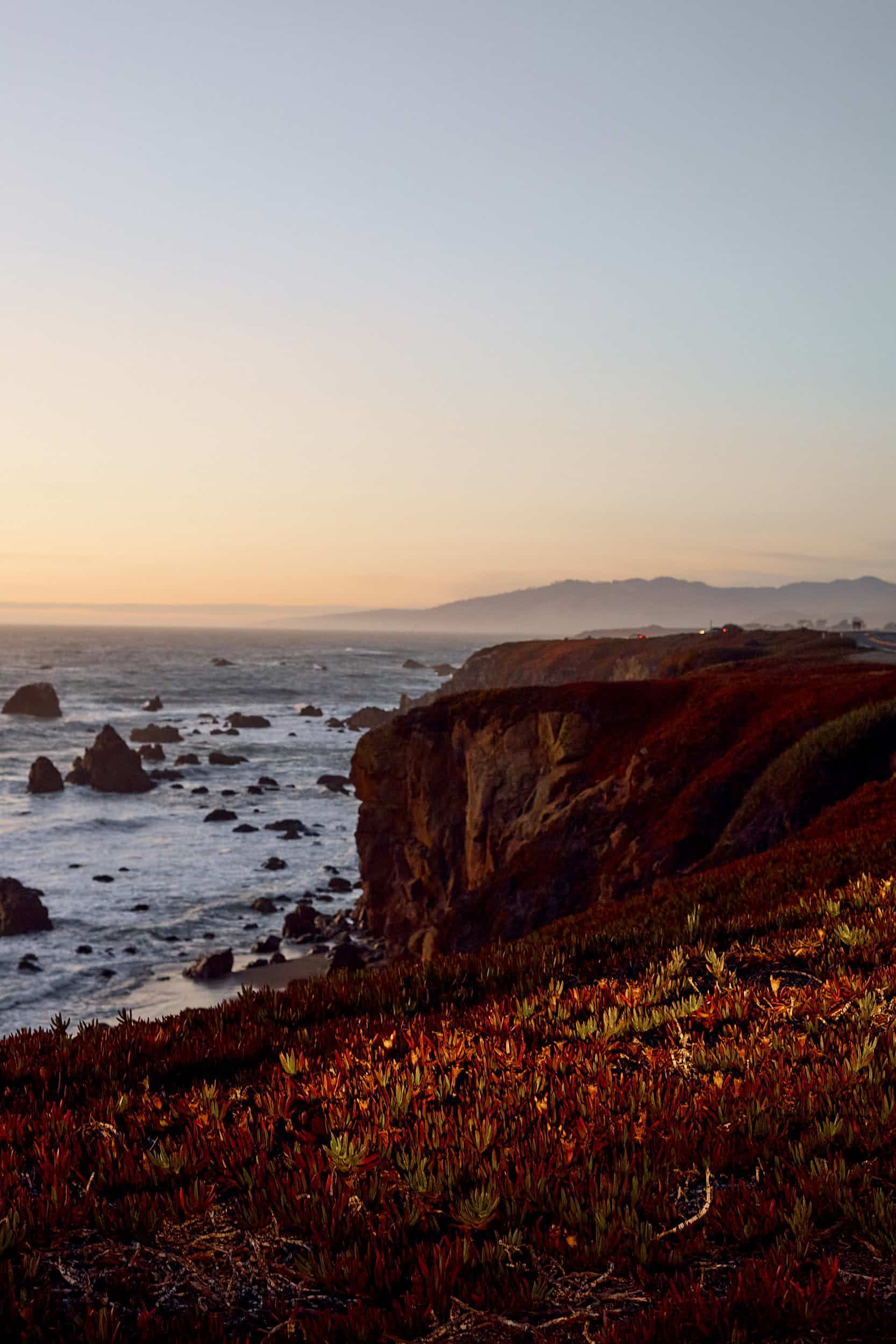 Coastal cliff at sunset, overlooking ocean and rocky shoreline. Warm colors illuminate vegetation and sky.