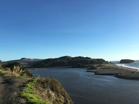Blue sky over a coastal scene with a river flowing into the ocean, hills in the distance.