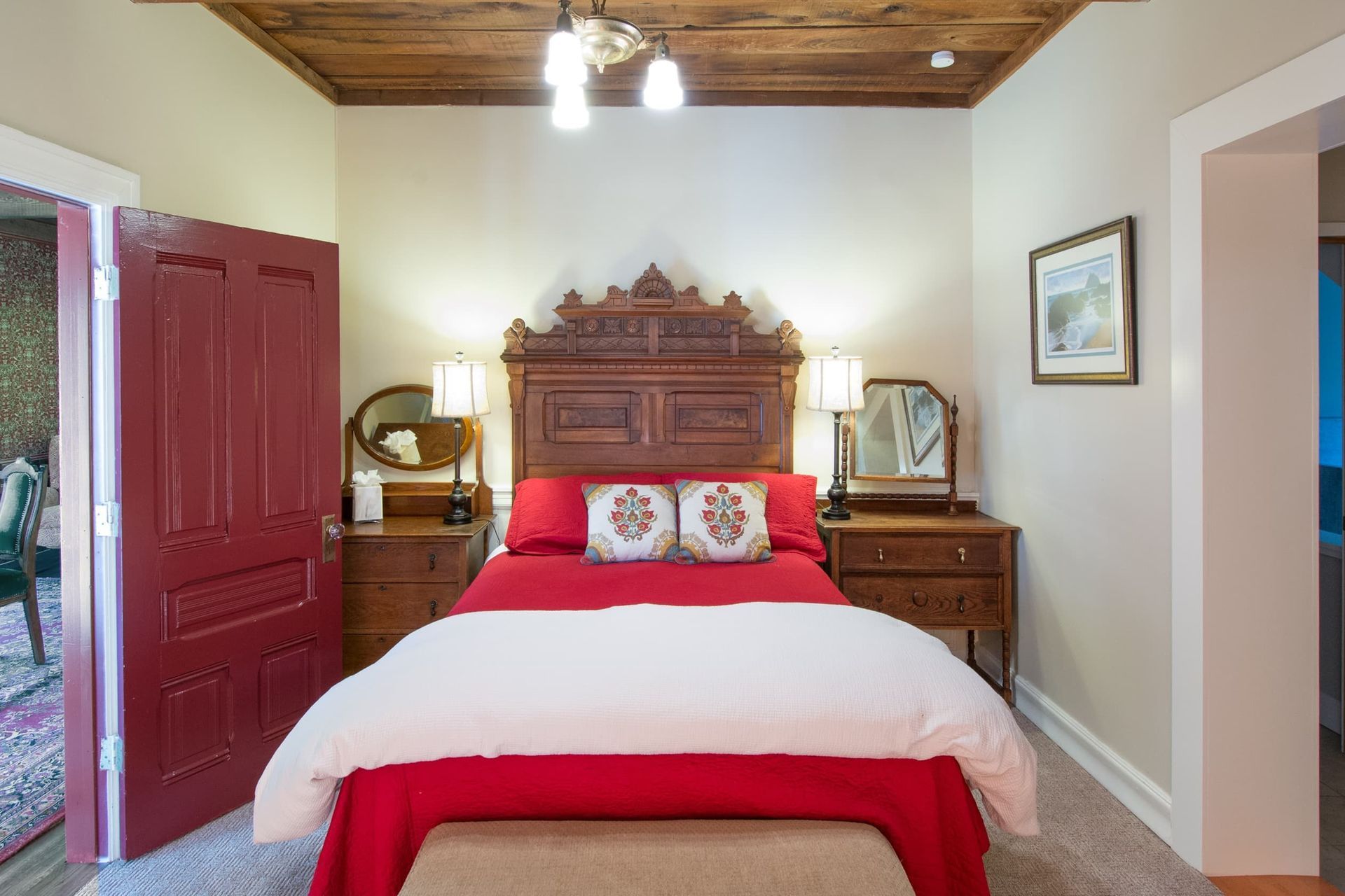 Bedroom with red bedding, dark wood headboard, bedside tables, red door, and exposed wood ceiling.