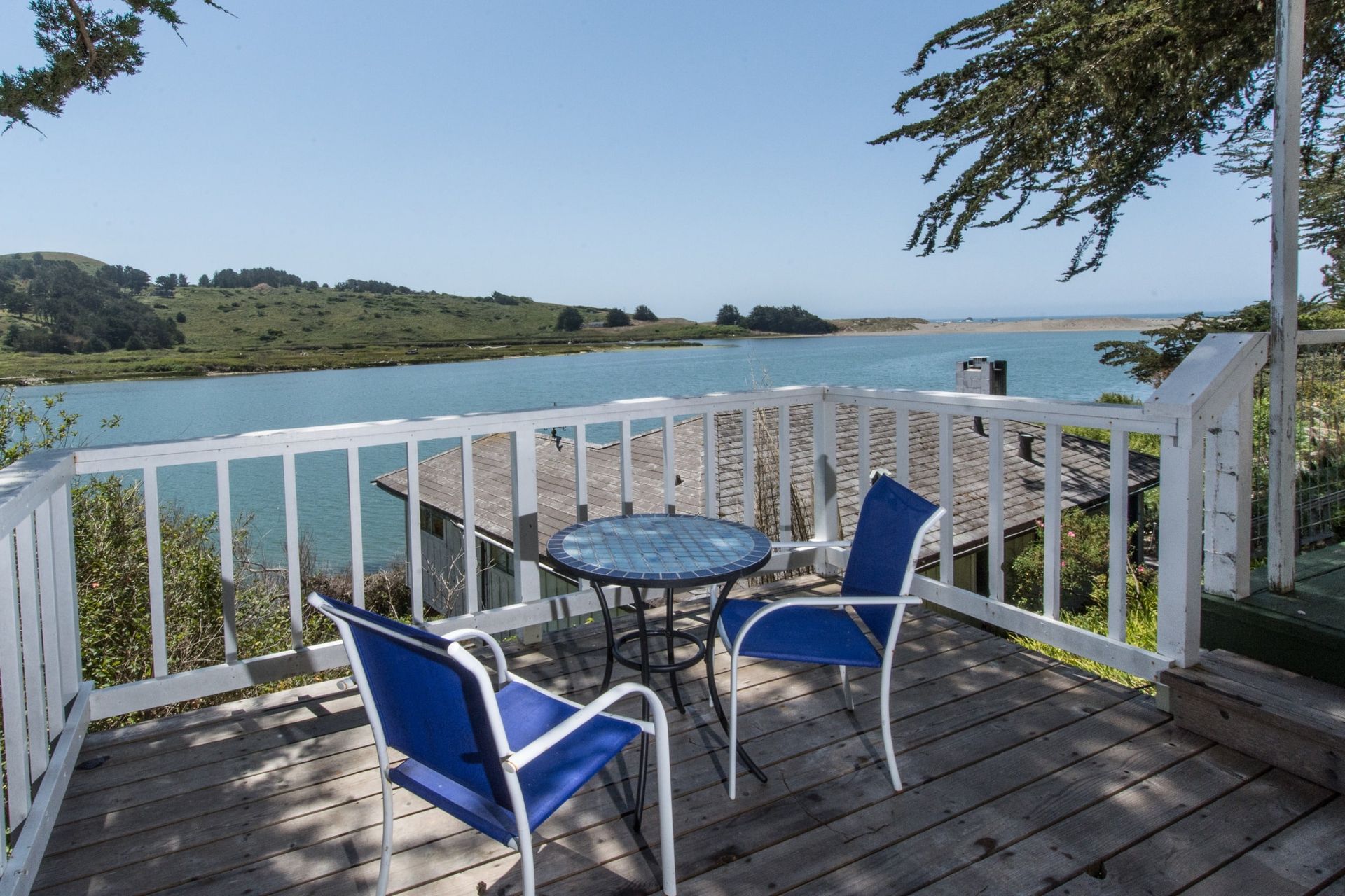 Deck with a table and two blue chairs overlooking a river on a sunny day.