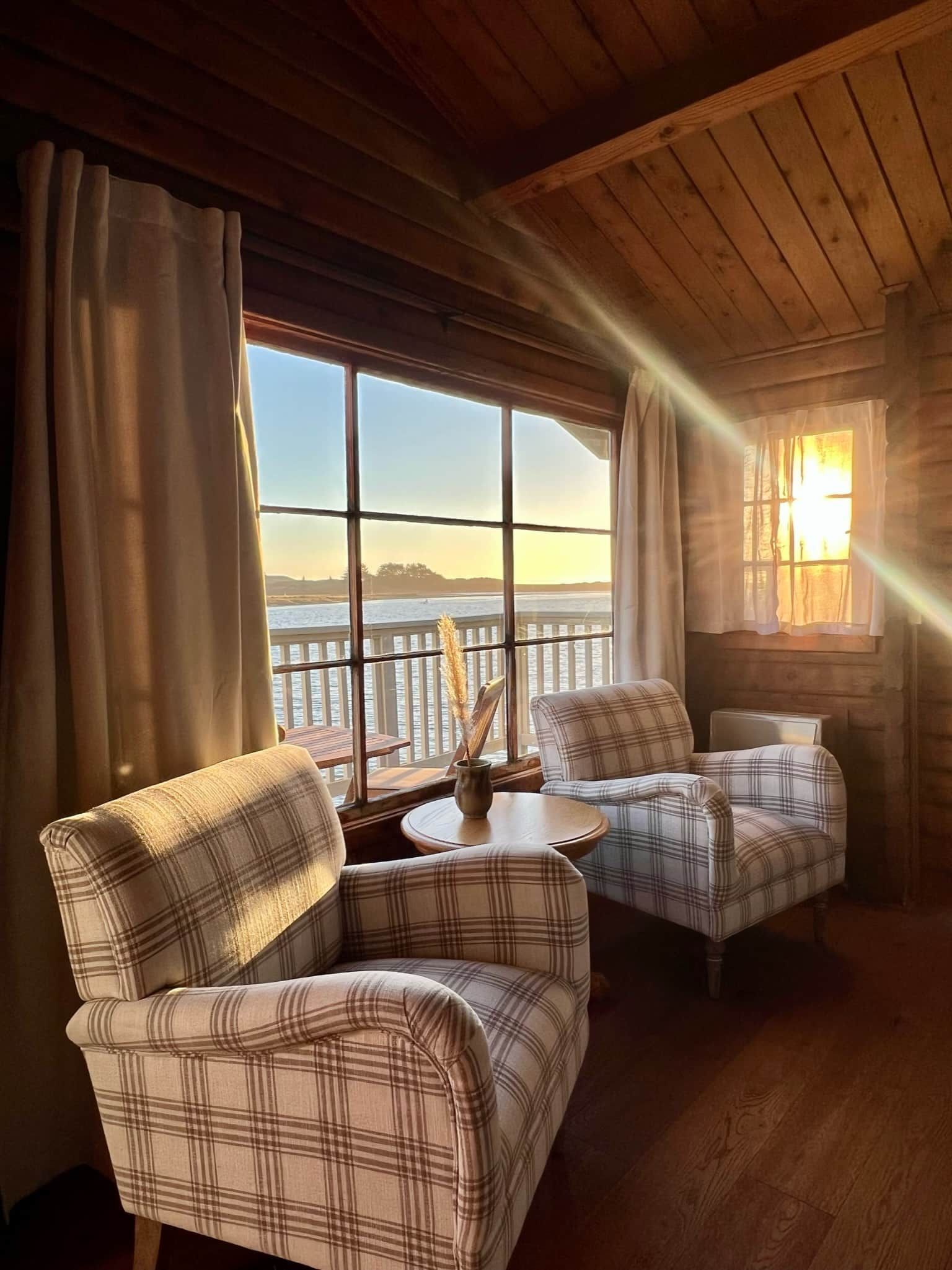 Cozy room with two patterned armchairs by a window overlooking a water view; sunlight streams through.
