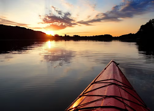 Kayak on calm water reflecting a sunset's orange and gold hues, silhouetted shoreline.