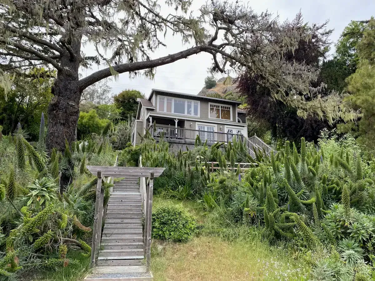 Wooden stairs lead up to a gray house nestled in lush greenery, under a cloudy sky.