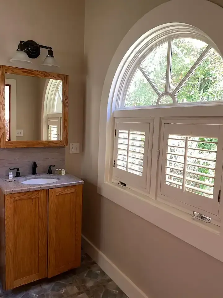Bathroom with wooden vanity, arched window with shutters, and wooden-framed mirror.