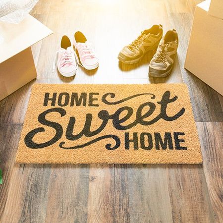 A welcome mat that says home sweet home with two pairs of shoes next to it and moving boxes