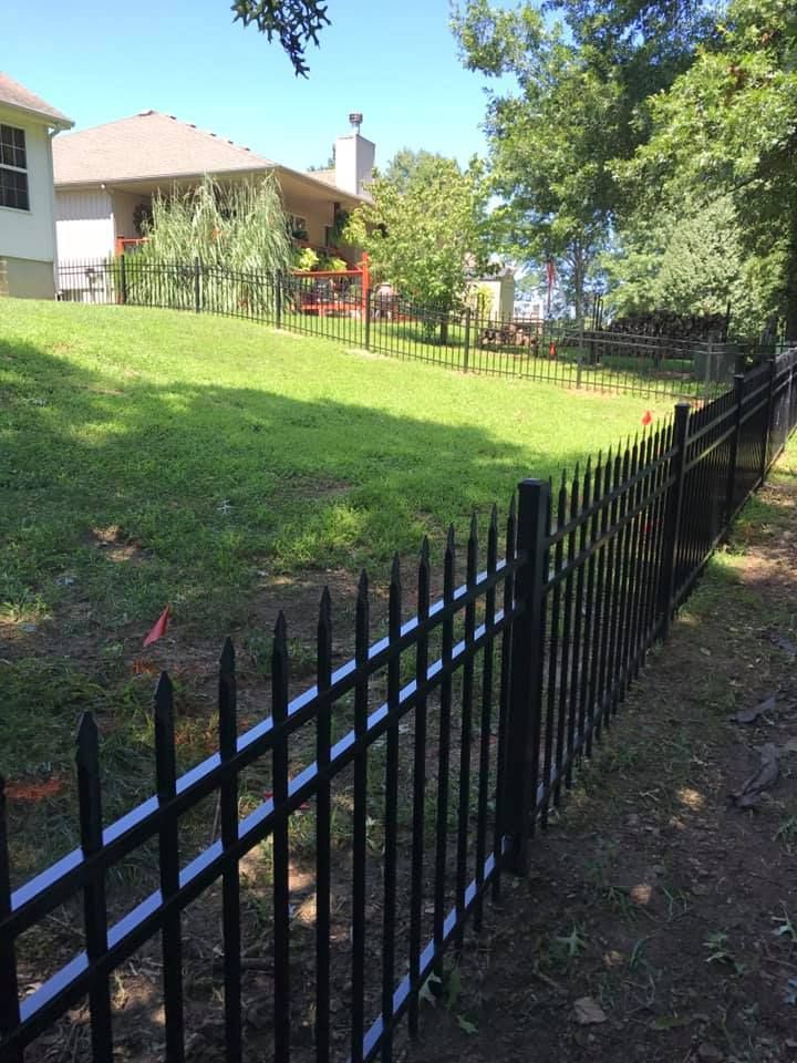 A black metal picket fence runs along a sloping grassy yard toward a house in the background on a sunny day.