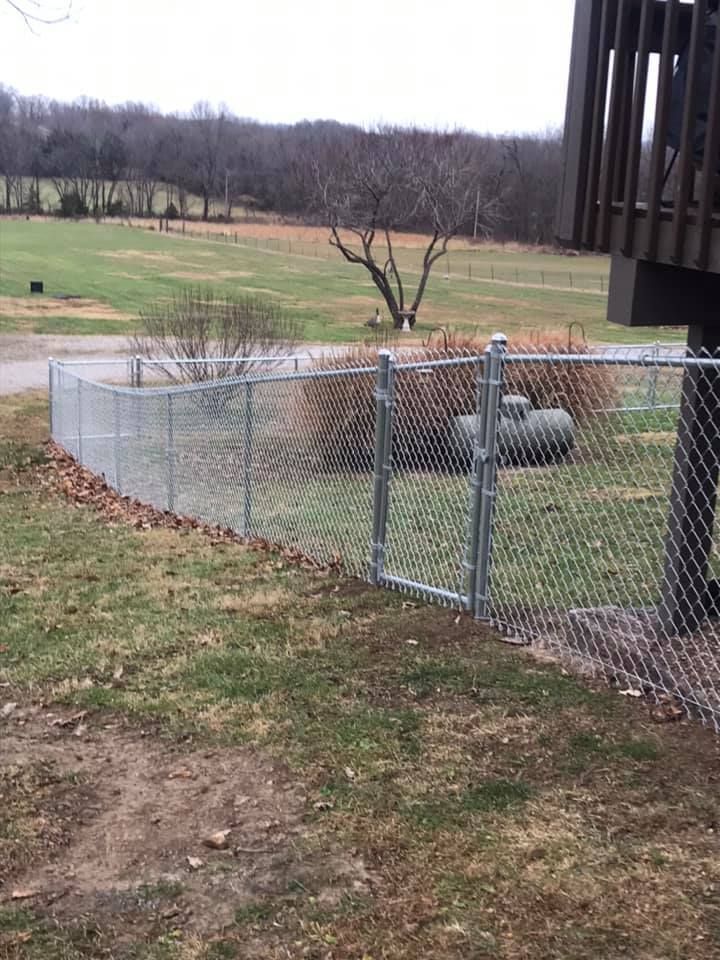 A chain-link fence with a closed gate runs across a grassy yard near a wooden deck, with a field and trees in the back.