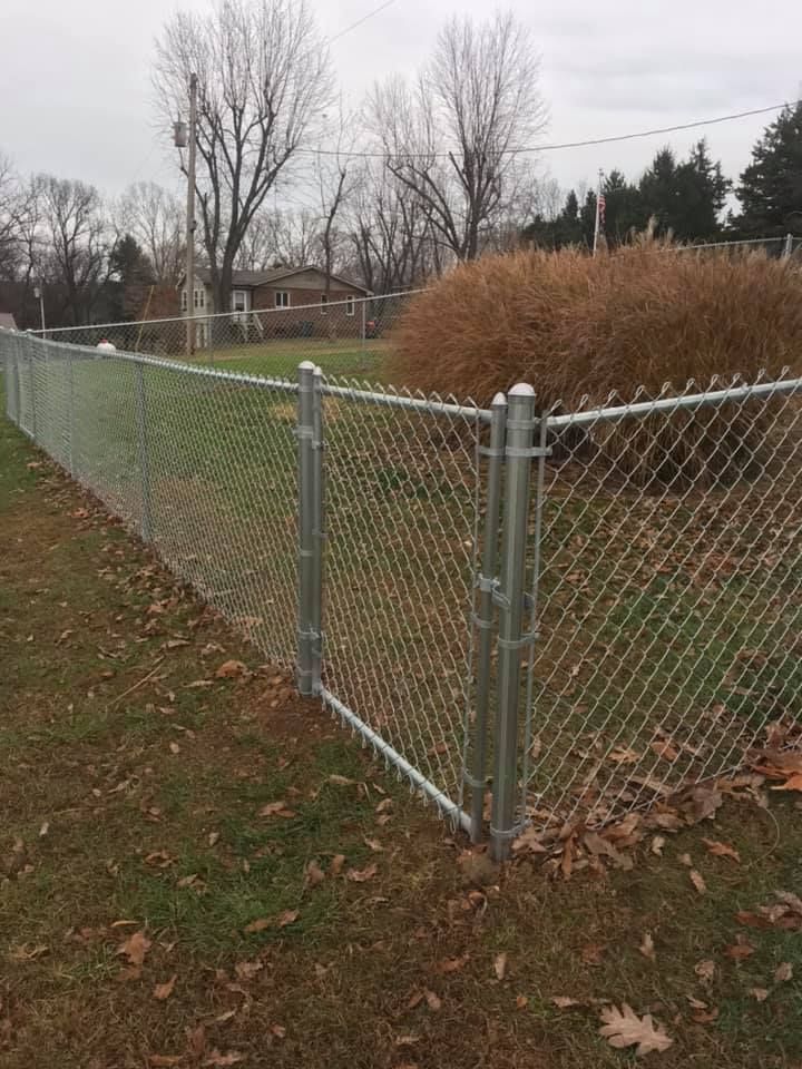 A chain-link fence with a gate forms a corner in a grassy yard, with a large bush and trees in the background.