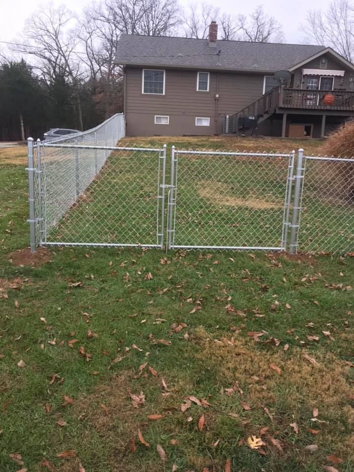 A double-gate chain-link fence stands in a grassy yard in front of a brown suburban house with a deck.