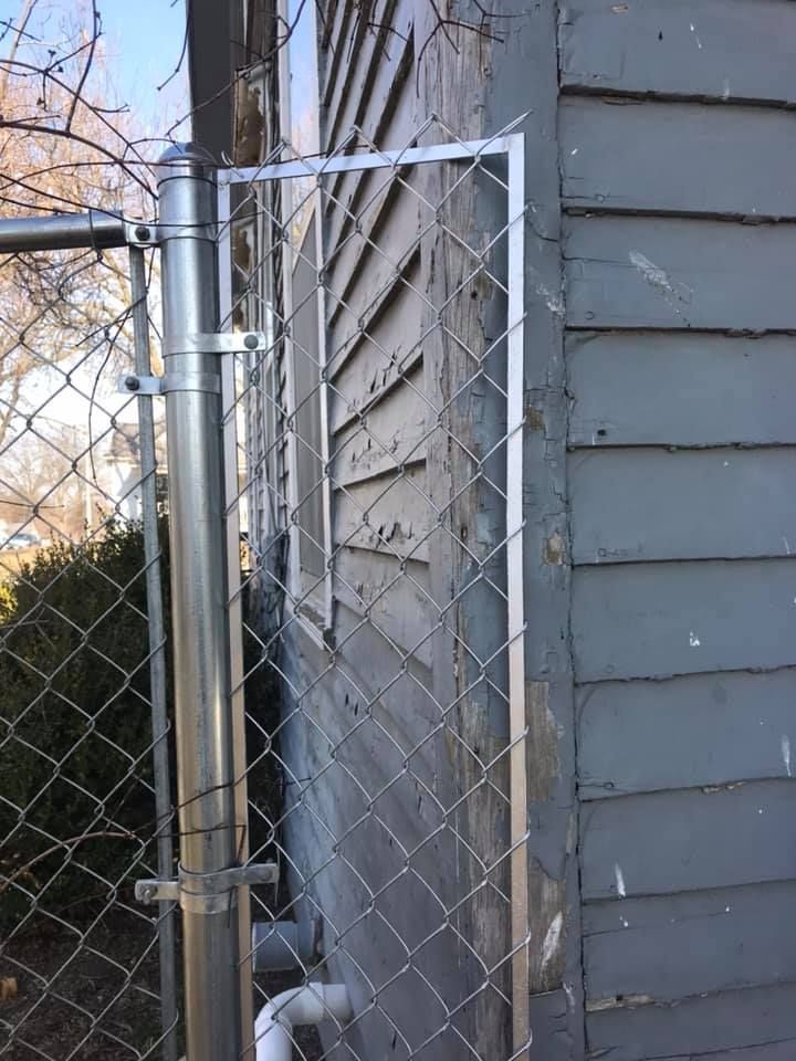 A chain-link fence attaches to the side of a gray, wood-sided house with a narrow metal frame filling the gap.