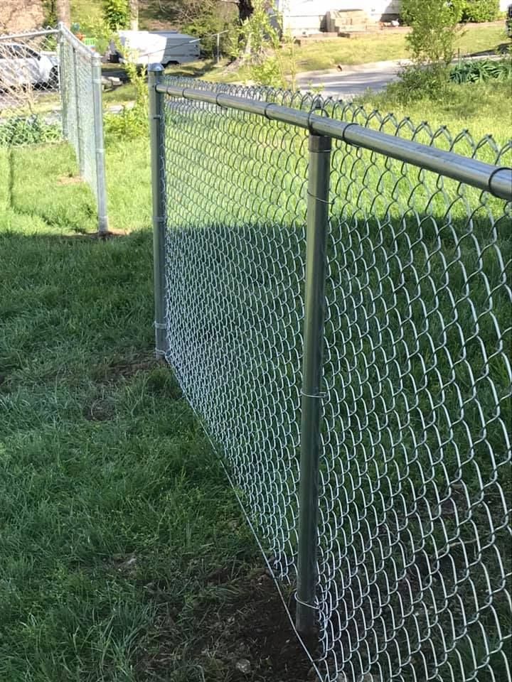 A silver chain-link fence stands on a grassy lawn next to a road, with another fence section visible in the background.
