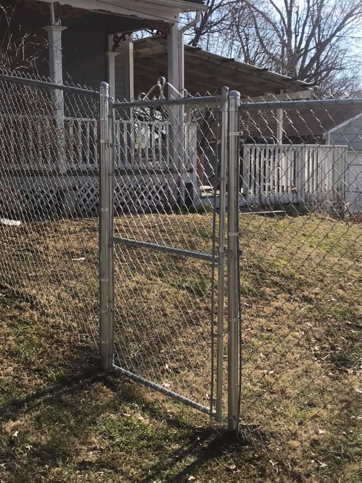 A metal chain-link fence gate stands in a residential yard with a house and porch in the background.
