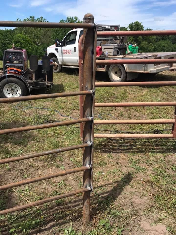 Welded metal fence rails attached to a vertical post in a grassy field with a truck and welding equipment in the background.