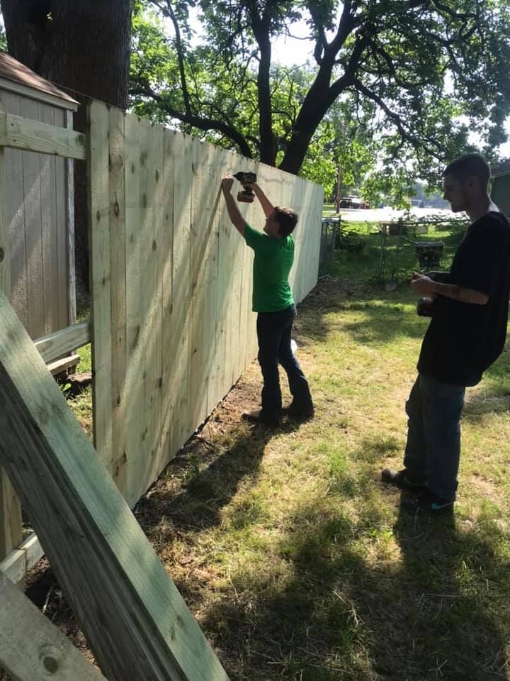 Two people working on a wooden fence outdoors, one person using a drill to attach a board.