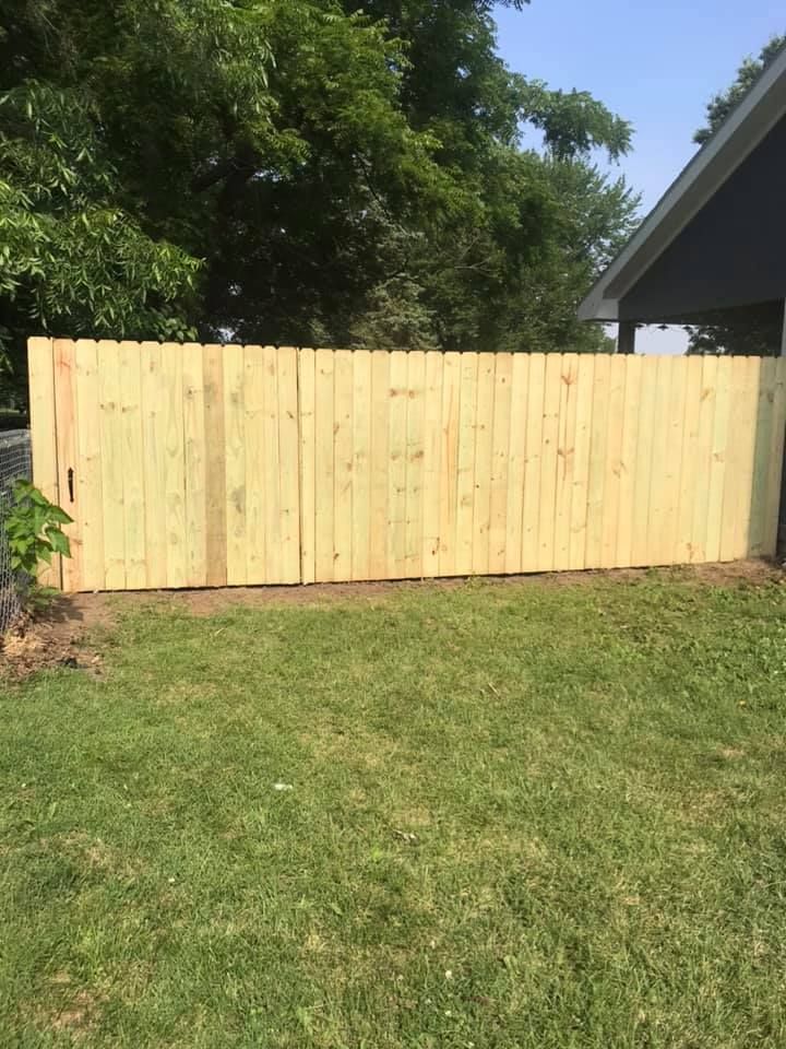 A newly installed wooden privacy fence stands in a grassy yard, partially shaded by trees next to a house roof.