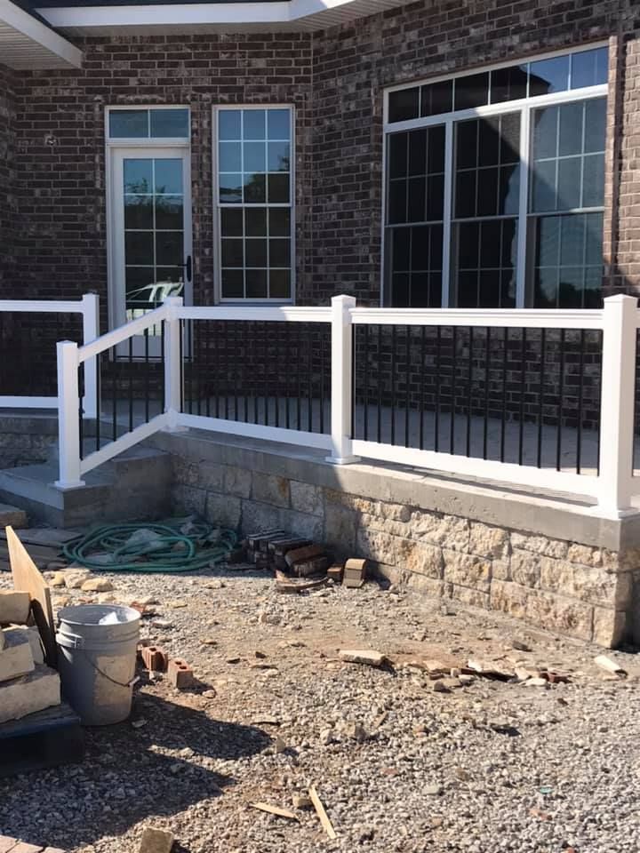 A brick house patio with white railings, black balusters, and a stone base under construction on a gravel ground.