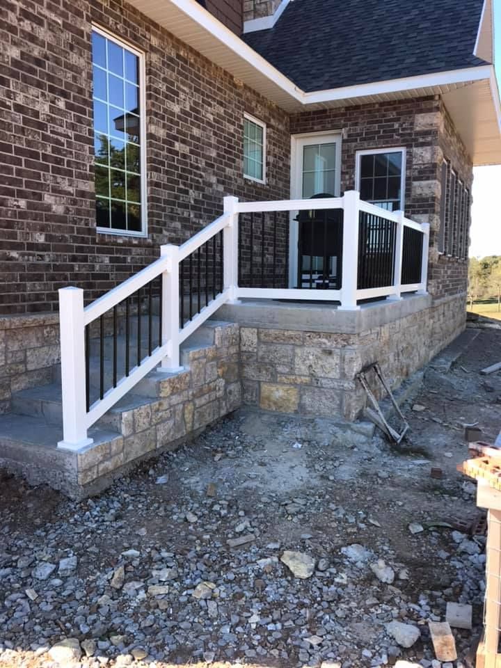 Exterior view of a stone house with a small concrete porch, a white railing, and stairs leading down to rocky ground.