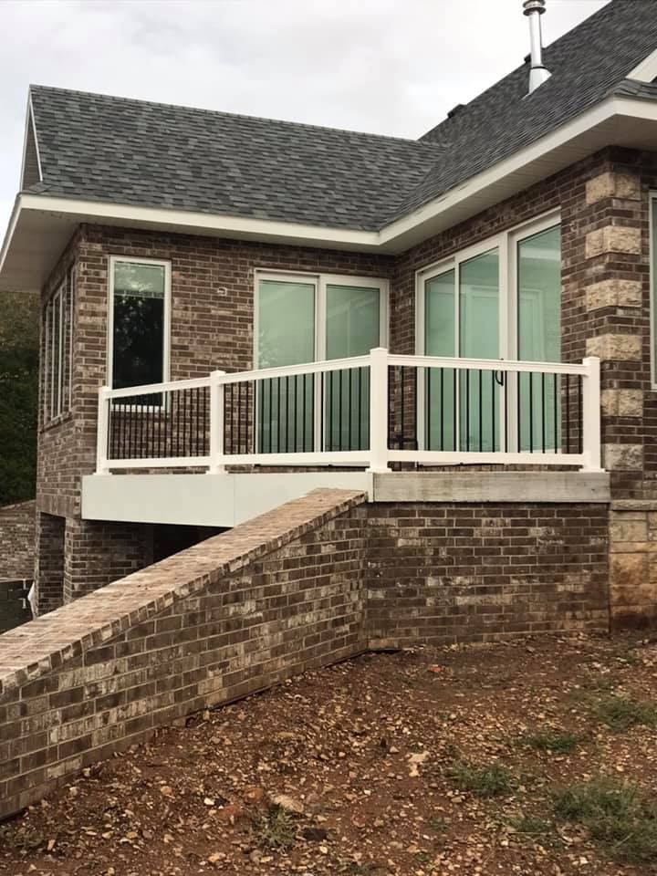 A brick house exterior with a white deck railing, large windows, and a sloped brick garden wall in the foreground.