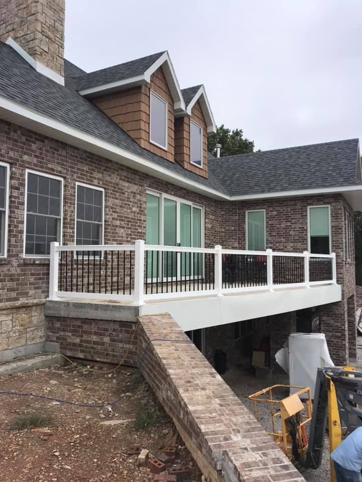 A house under construction with a brick exterior, shingled roof, dormer windows, and a raised white deck railing.