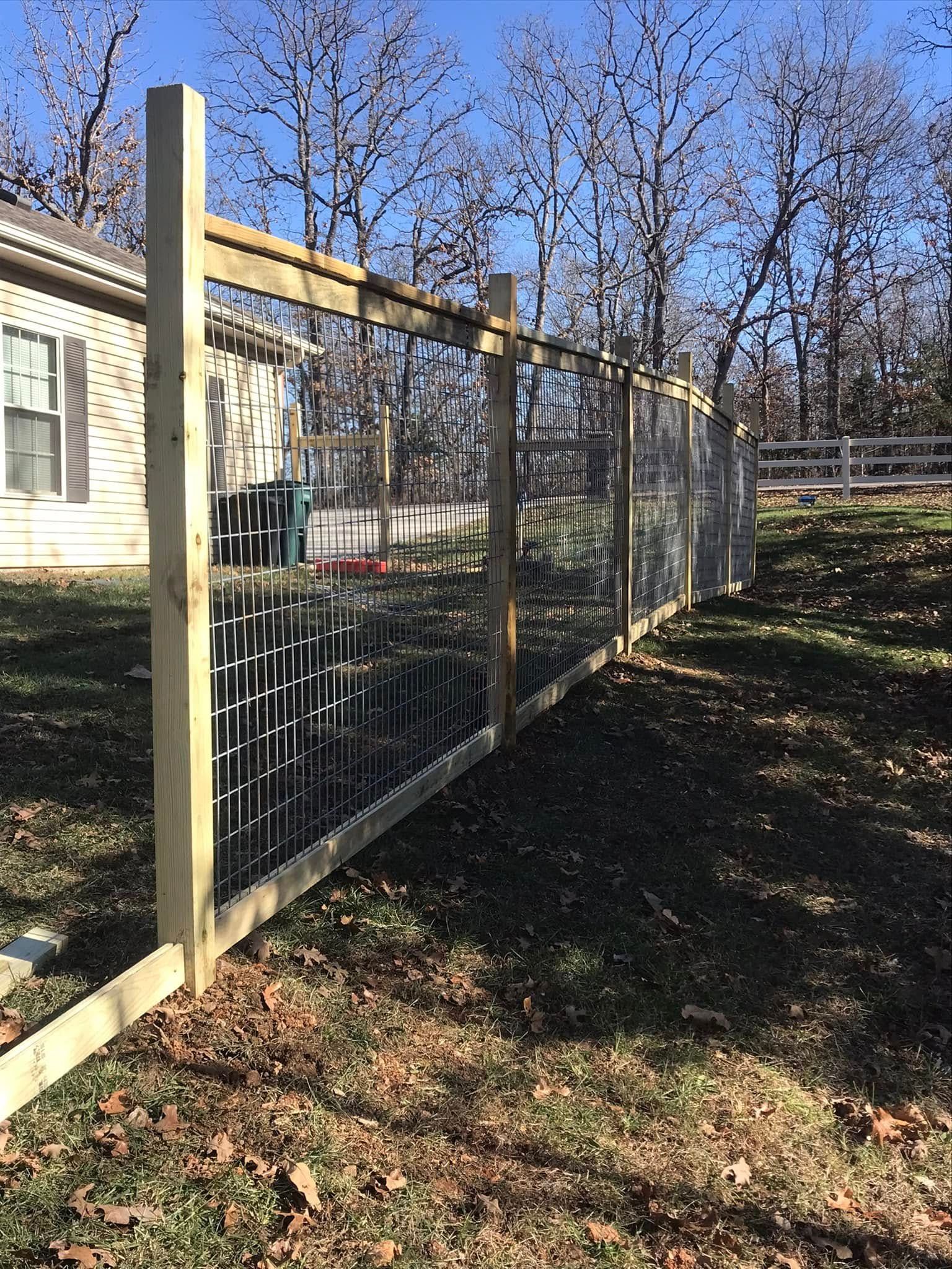 A partially constructed wooden fence with metal mesh wire panels stands in a grassy yard under a clear blue sky.